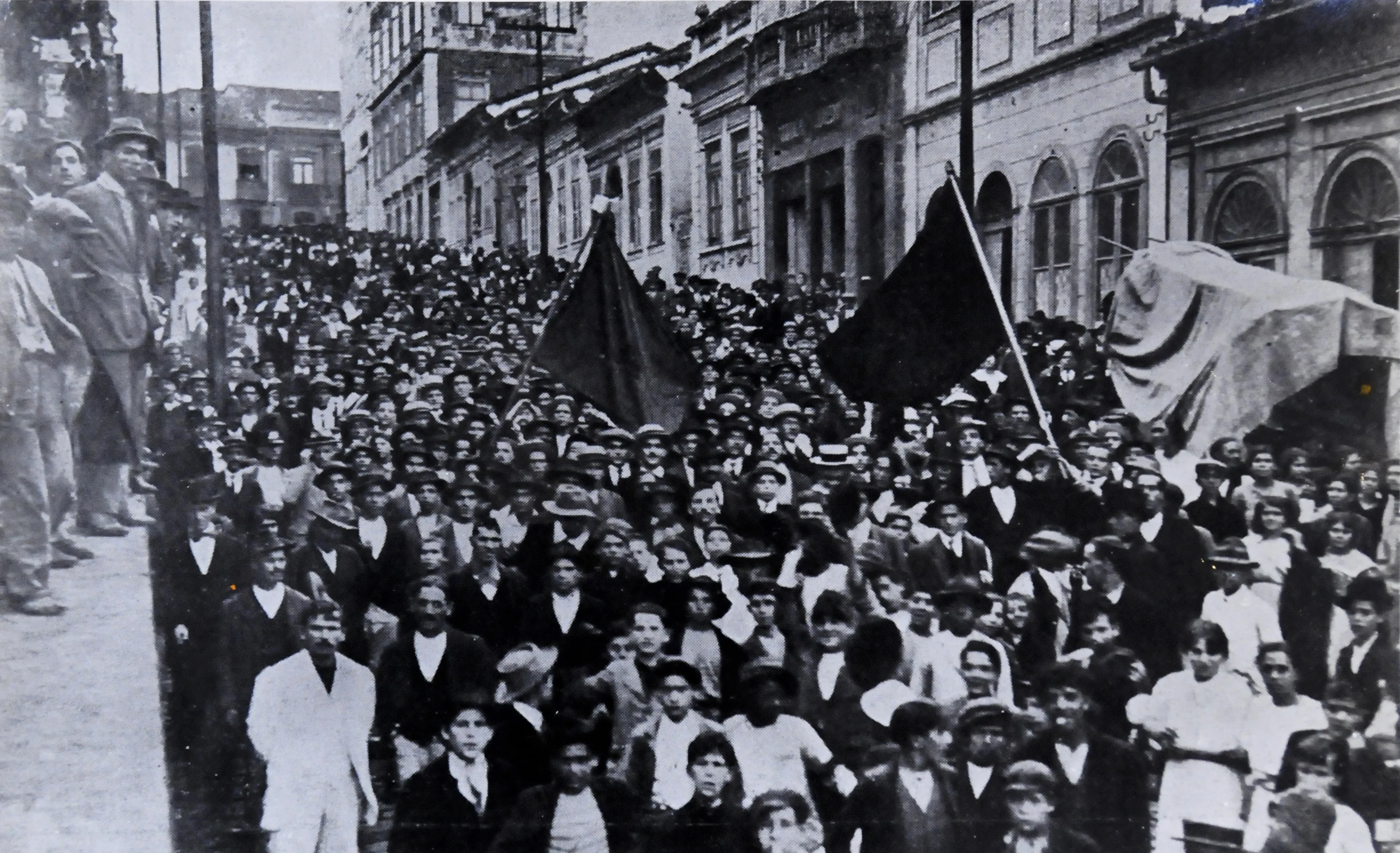 Fotografia histórica da Greve Geral de 1917 com multidão de trabalhadores descendo rua de paralelepípedos empunhando bandeiras negras.