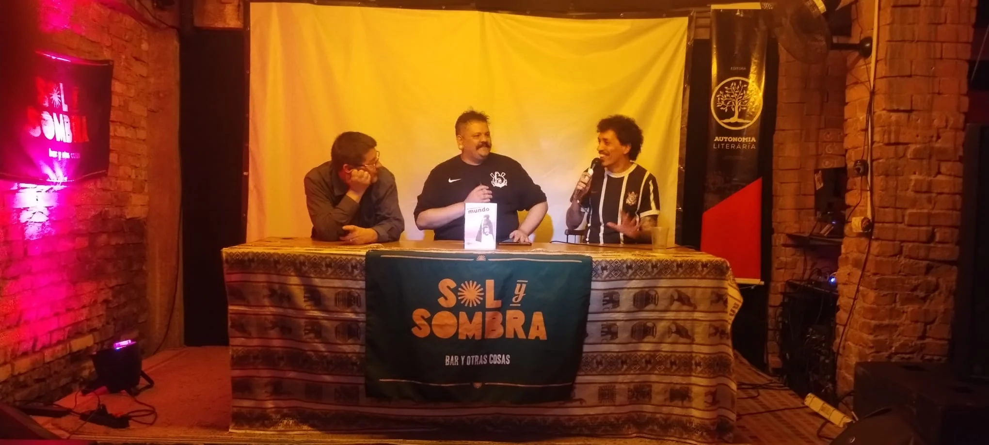 Três homens sentados a uma mesa de debate com toalha escrita 'Sol y Sombra' e exemplar do livro 'Da rodoviária para o mundo' ao centro, com o homem à direita falando ao microfone e banner da Autonomia Literária ao fundo.