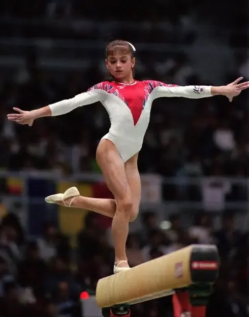 Dominique Moceanu of ‘Magnificent Seven’ wants to rebuild the culture of gymnastics A ginasta Dominique Moceanu em collant branco e vermelho equilibrando-se sobre uma perna na trave de equilíbrio durante apresentação olímpica.