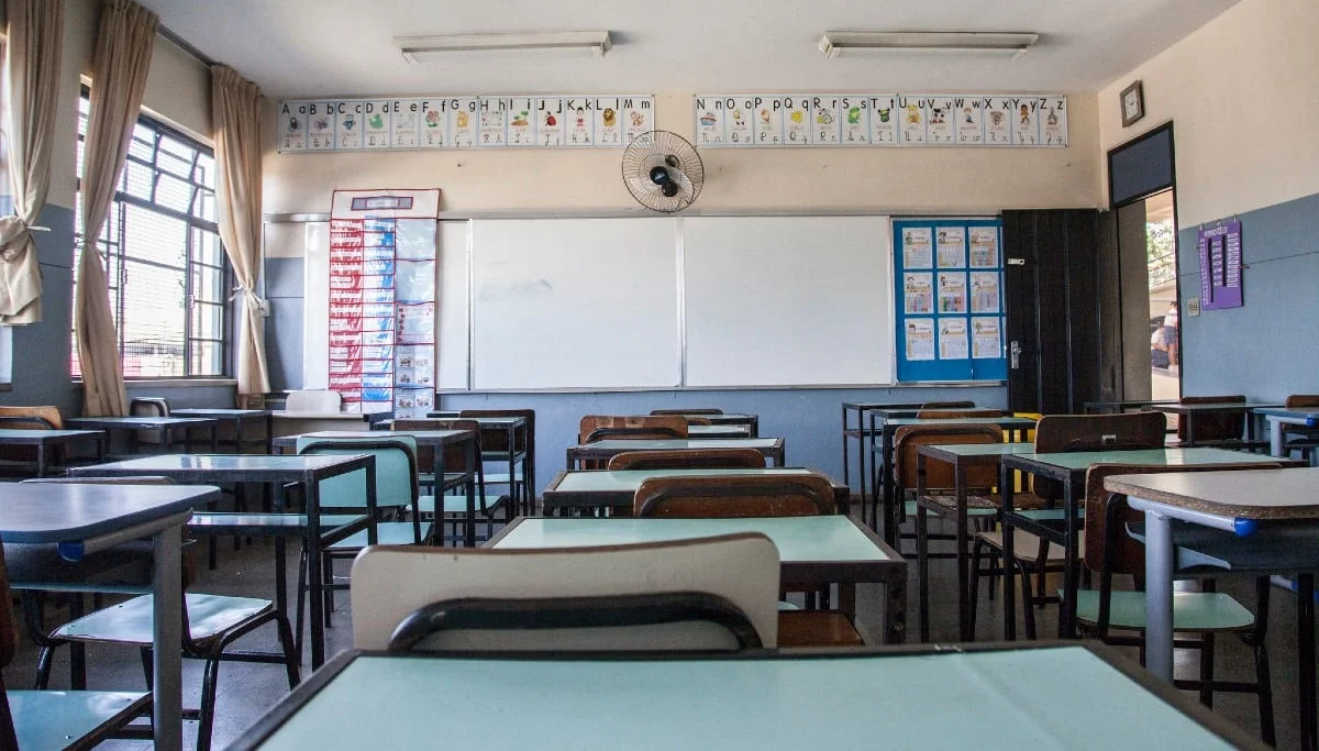 Sala de aula de escola pública vazia, com fileiras de carteiras escolares de tampo verde, lousa branca e ventilador de parede.