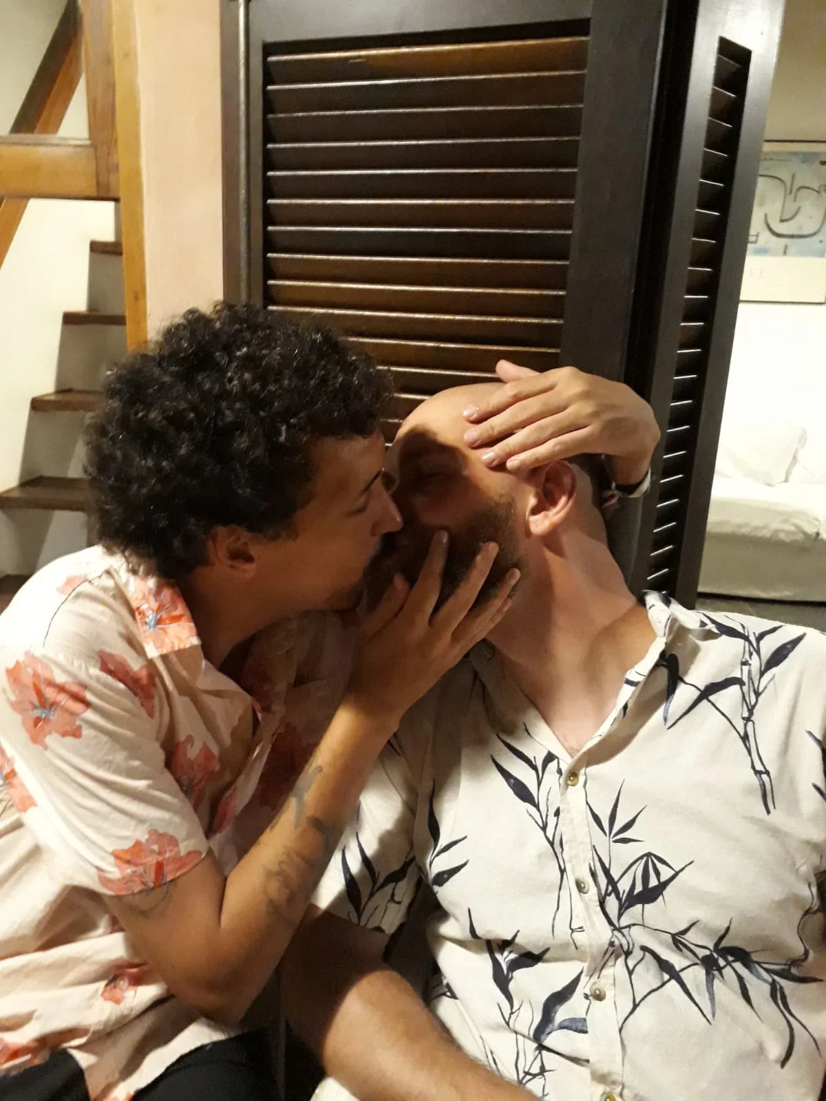 Homem de camisa floral beijando carinhosamente no rosto outro homem de barba e cabeça raspada.