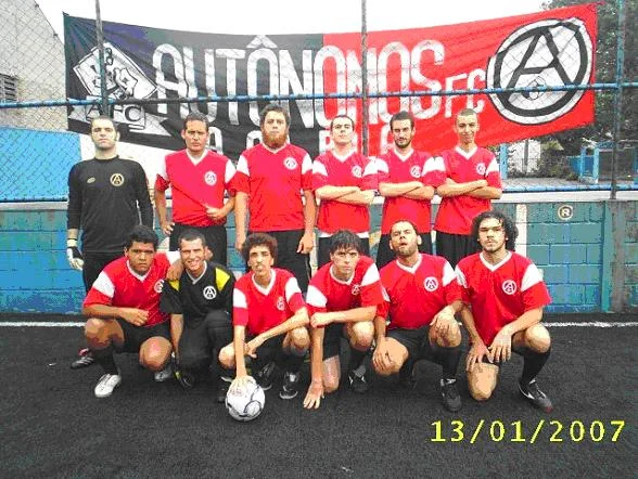 Time de futebol amador Autônomos FC perfilado em quadra com uniforme vermelho e preto, posando em frente a faixa do time com símbolo anarquista e a data 13/01/2007.