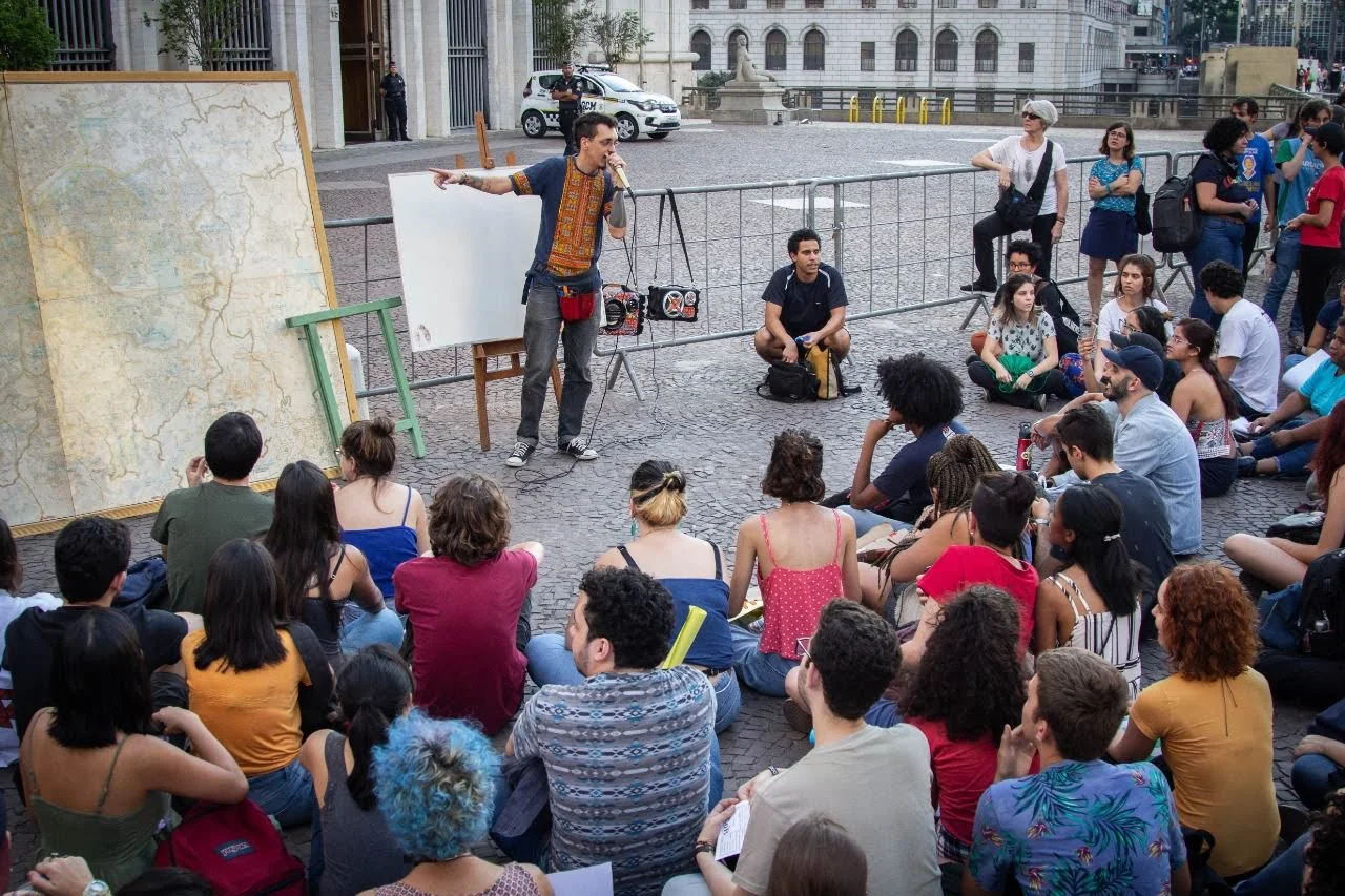 Homem em pé falando ao microfone diante de quadro branco e cavalete com mapa, em aula pública para dezenas de estudantes sentados no chão de praça pública.