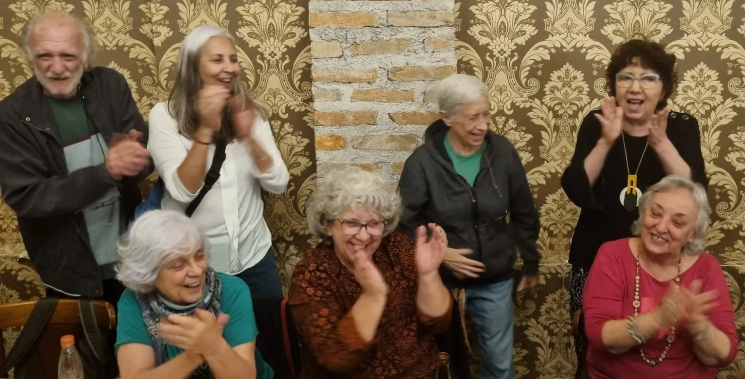 Oito pessoas idosas sorridentes aplaudindo diante de parede com papel de parede dourado e pilar de tijolos aparentes.