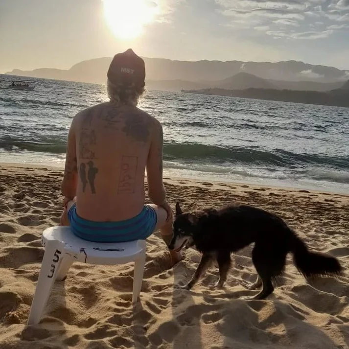 Pessoa sem camisa de costas sentada em banquinho plástico na praia ao entardecer, de boné do MST, observando o mar ao lado de um cachorro preto na areia.