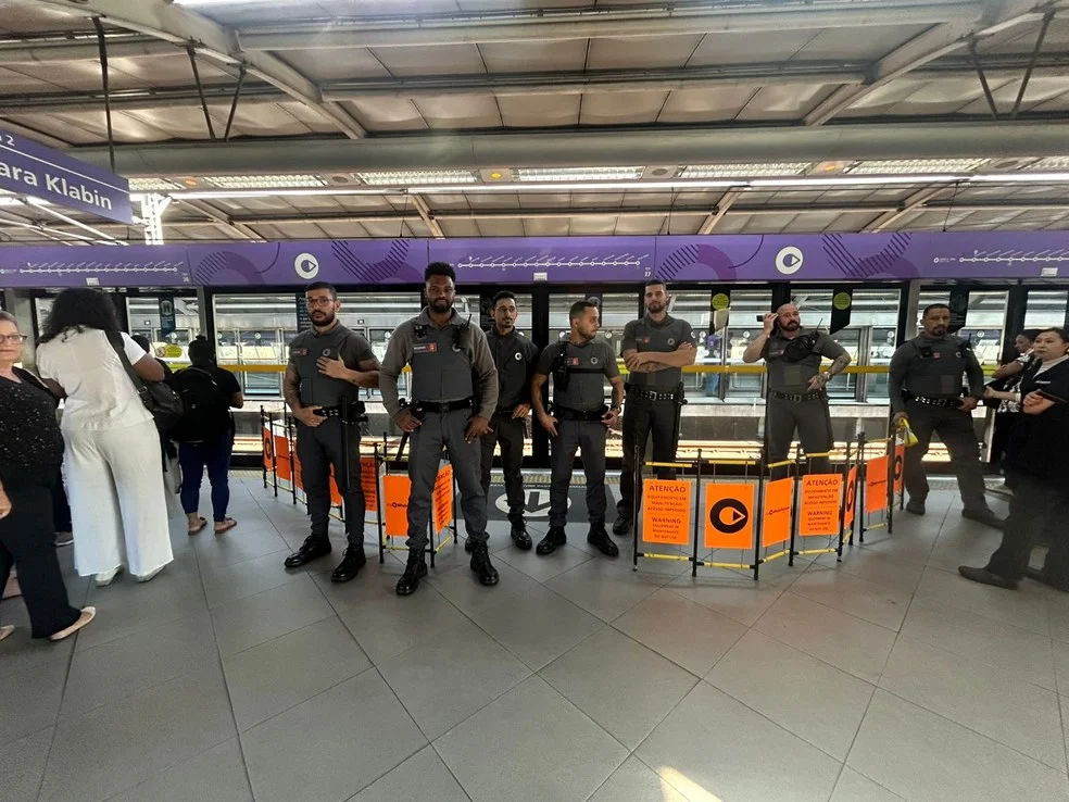 Porta de plataforma fechada na estação Campo Limpo, sentido Chácara Klabin após acidente — Foto: Beatriz Backes/TV Globo Sete agentes de segurança uniformizados alinhados em frente às portas de plataforma de estação sobre trilhos com cavaletes de aviso 'Atenção'.