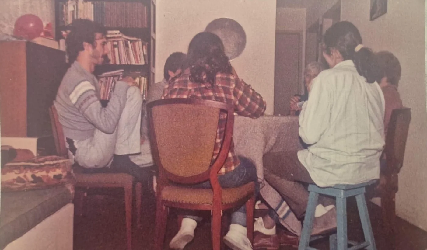 Fotografia antiga de pessoas reunidas ao redor de uma mesa jogando cartas (jogo de buraco), em sala com estante de livros ao fundo.