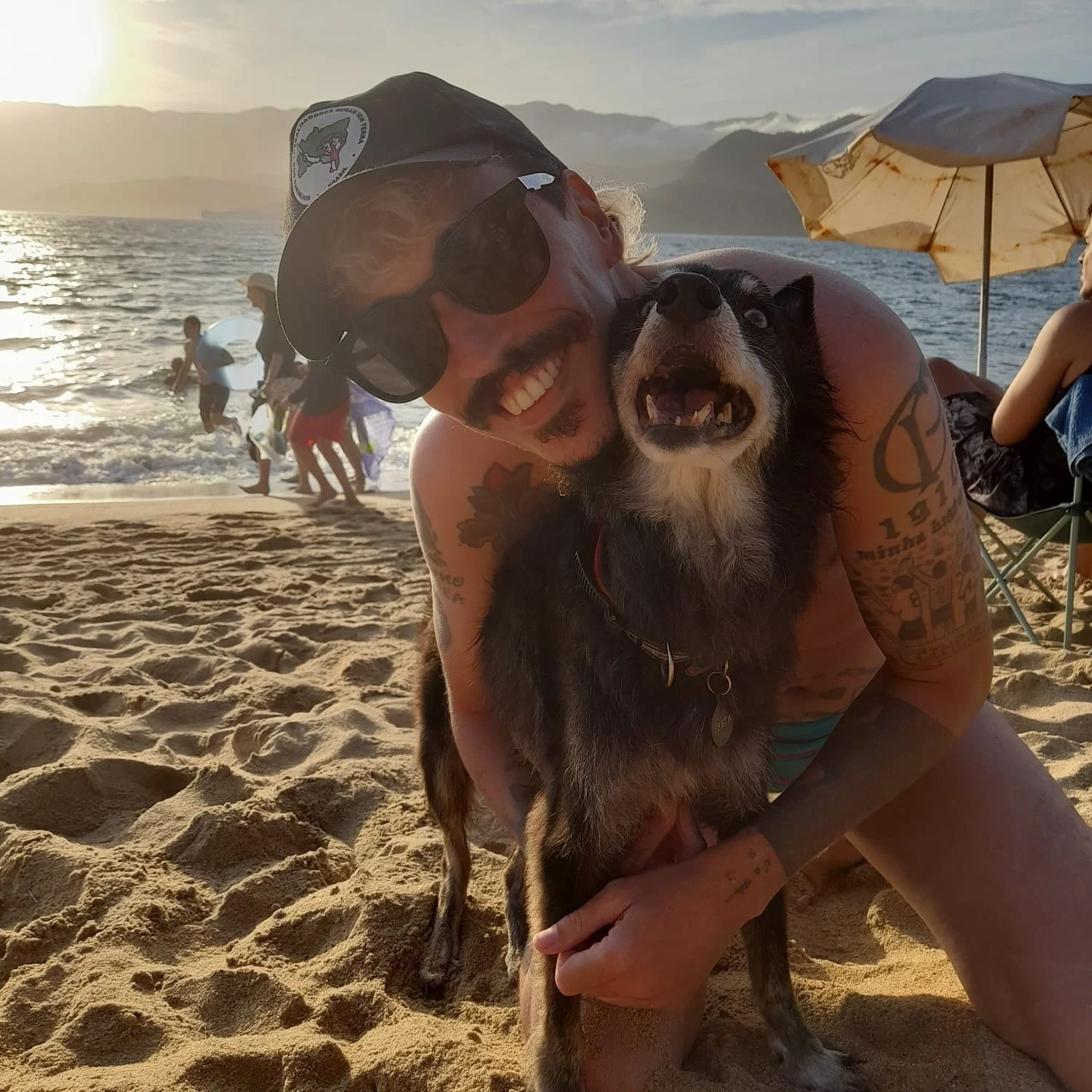 Homem sorrindo na praia, de óculos escuros e boné do MST, abraçado a uma cadela preta na areia, com banhistas e o mar ao fundo.